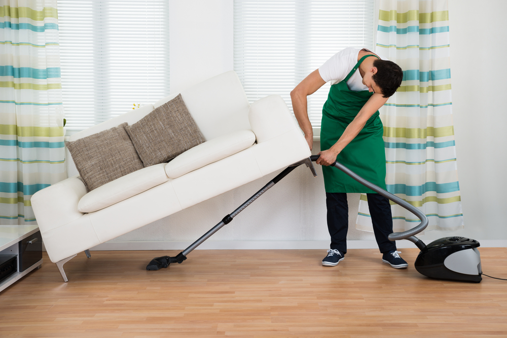 seorang pria sedang mengangkat sofa sembari membersihkan permukaan lantai di bawah sofa dengan vacuum cleaner