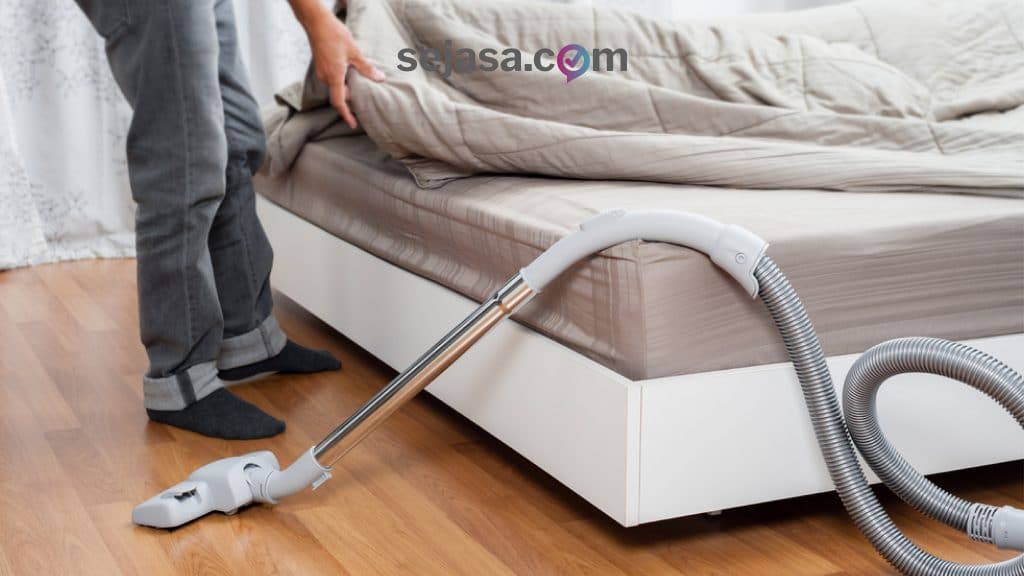 seseorang sedang melakukan vakum lantai sembari membereskan tempat tidurnya demi menjaga kebersihan rumah